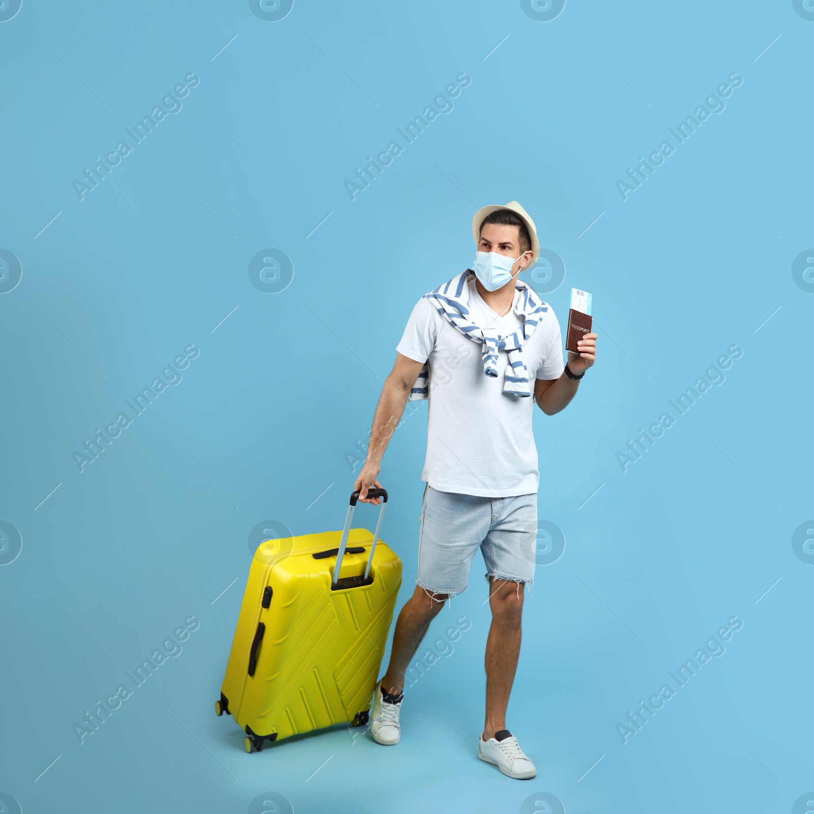 Male tourist in protective mask holding passport with ticket and suitcase on turquoise background Photo of Male tourist in protective mask holding passport with ticket and suitcase on turquoise background