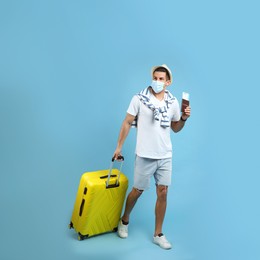 Male tourist in protective mask holding passport with ticket and suitcase on turquoise background Photo of Male tourist in protective mask holding passport with ticket and suitcase on turquoise background