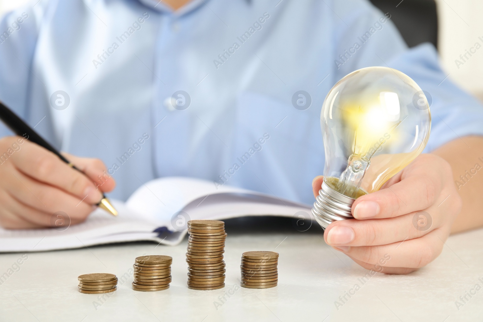 Woman with light bulb, notebook and coins at white table, closeup. Power saving Photo of Woman with light bulb, notebook and coins at white table, closeup. Power saving