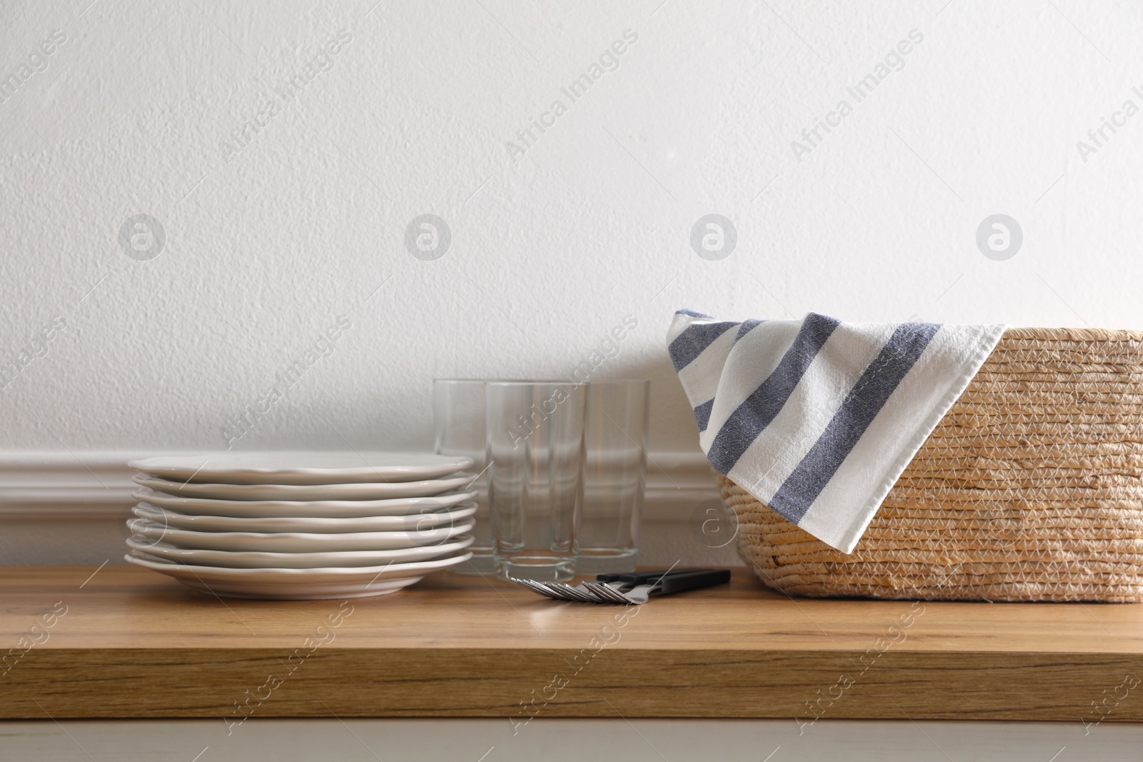Kitchen towel in wicker basket and clean dishware on wooden table near white wall Photo of Kitchen towel in wicker basket and clean dishware on wooden table near white wall
