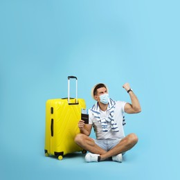 Male tourist in protective mask holding passport with ticket and suitcase on turquoise background, space for text Photo of Male tourist in protective mask holding passport with ticket and suitcase on turquoise background, space for text