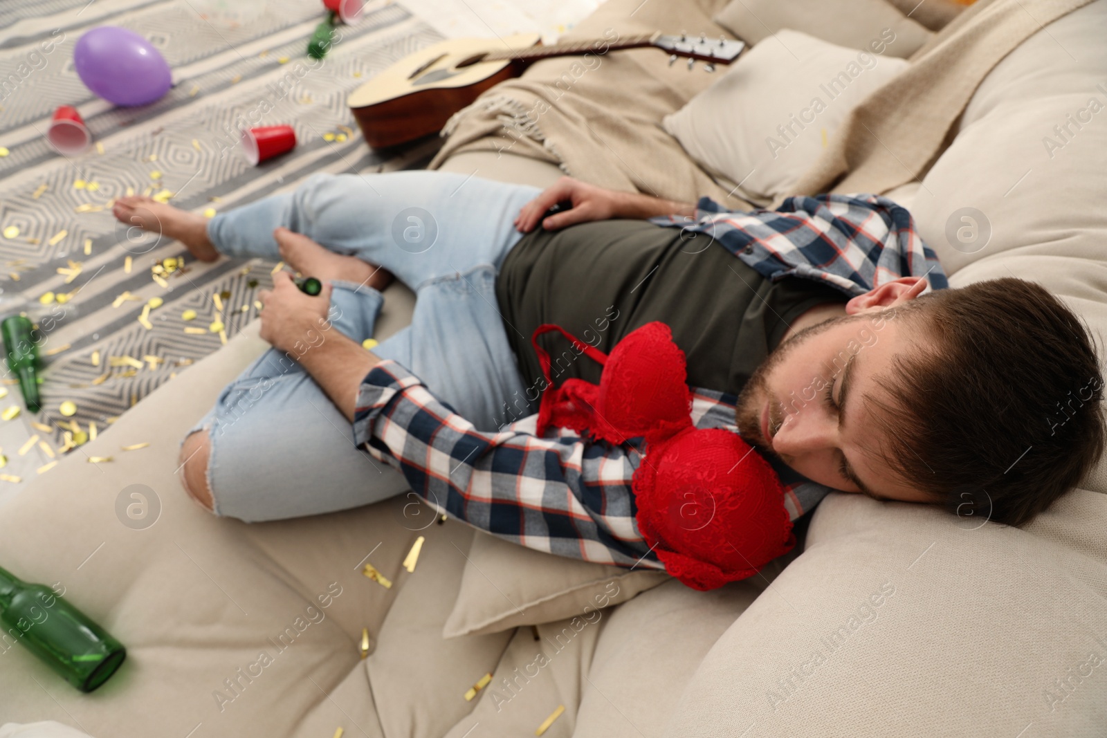 Young man with red bra sleeping on sofa in messy room after party Photo of Young man with red bra sleeping on sofa in messy room after party