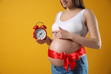 Young pregnant woman with alarm clock and red bow on yellow background, closeup. Time to give birth Photo of Young pregnant woman with alarm clock and red bow on yellow background, closeup. Time to give birth