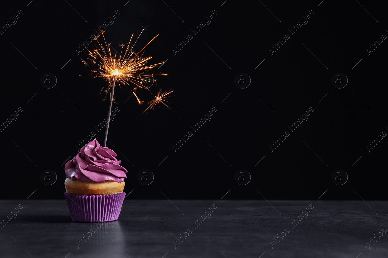 Delicious birthday cupcake with sparkler on table against black background Photo of Delicious birthday cupcake with sparkler on table against black background