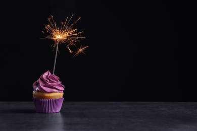 Delicious birthday cupcake with sparkler on table against black background Photo of Delicious birthday cupcake with sparkler on table against black background