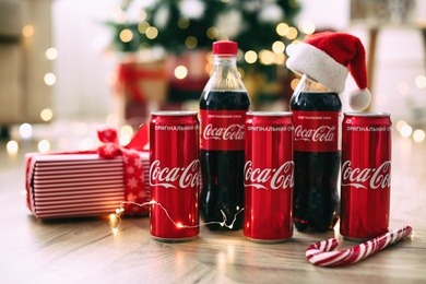 MYKOLAIV, UKRAINE - January 01, 2021: Bottles and cans of Coca-Cola on floor against blurred Christmas lights Photo of MYKOLAIV, UKRAINE - January 01, 2021: Bottles and cans of Coca-Cola on floor against blurred Christmas lights