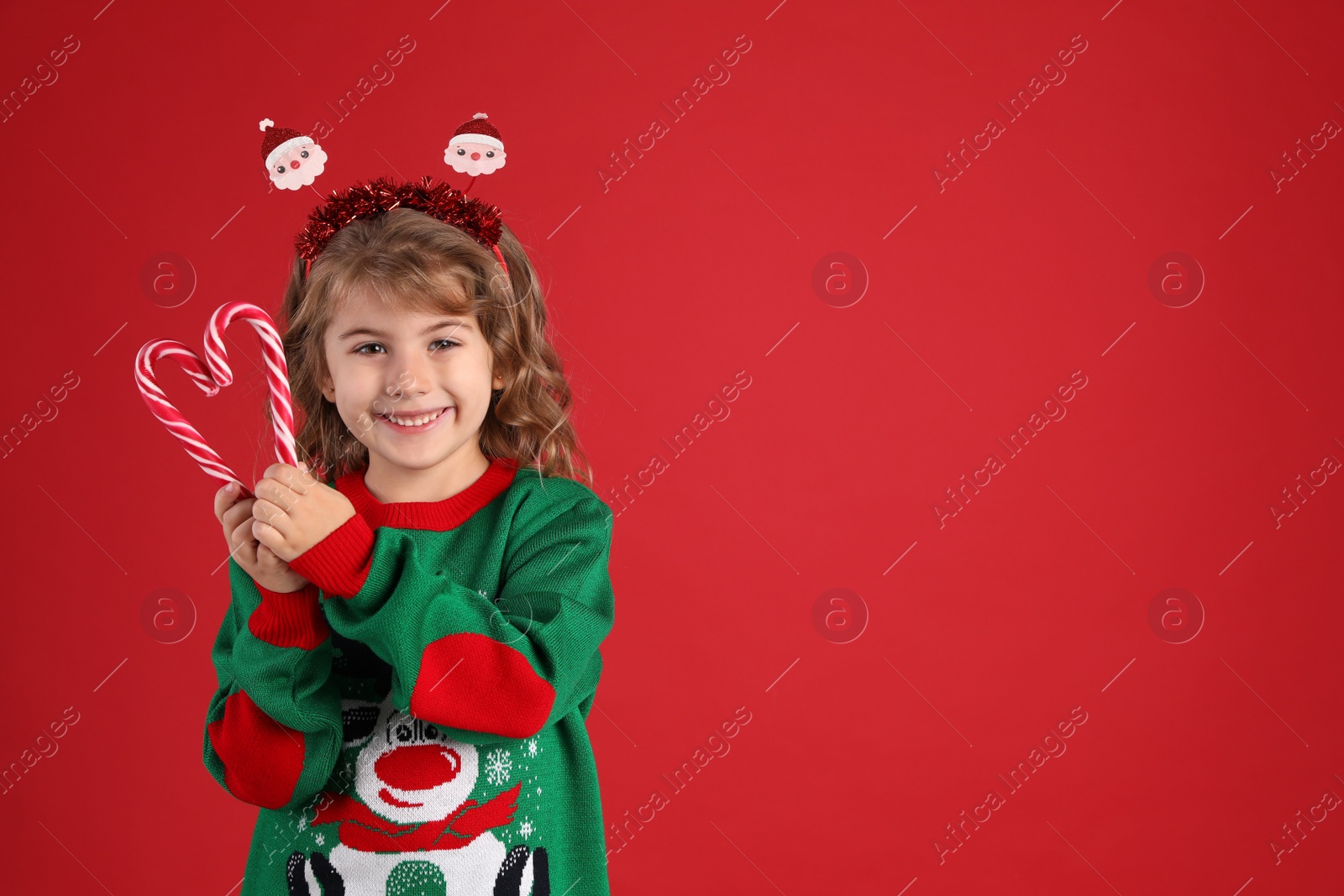 Cute little girl in Christmas sweater making heart shape with candy canes against red background. Space for text Photo of Cute little girl in Christmas sweater making heart shape with candy canes against red background. Space for text