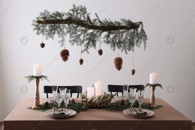 Photo of Christmas celebration. Cones hanging from fir tree branch over table with burning candles and tableware