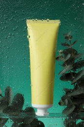 Tube with moisturizing cream and eucalyptus branches on green background, view through wet glass Photo of Tube with moisturizing cream and eucalyptus branches on green background, view through wet glass