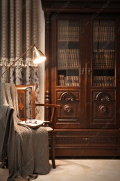 Comfortable armchair with book, blanket and lamp near wooden bookcase in library Photo of Comfortable armchair with book, blanket and lamp near wooden bookcase in library