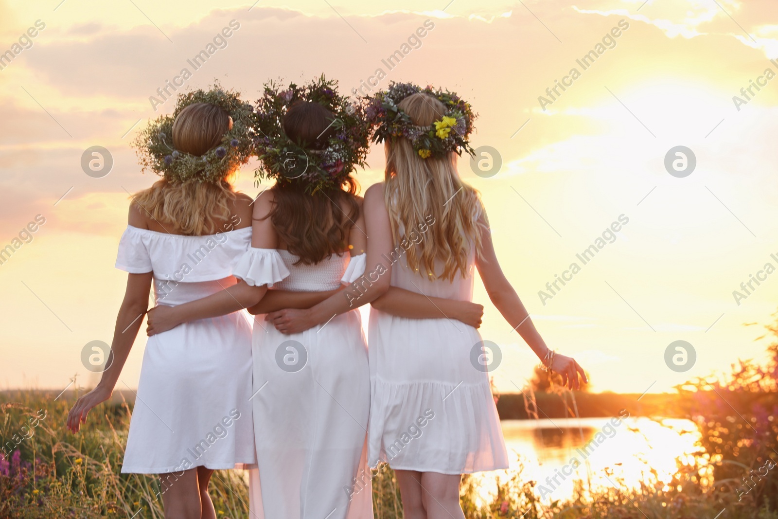 Young women wearing wreaths made of flowers outdoors at sunset, back view Photo of Young women wearing wreaths made of flowers outdoors at sunset, back view
