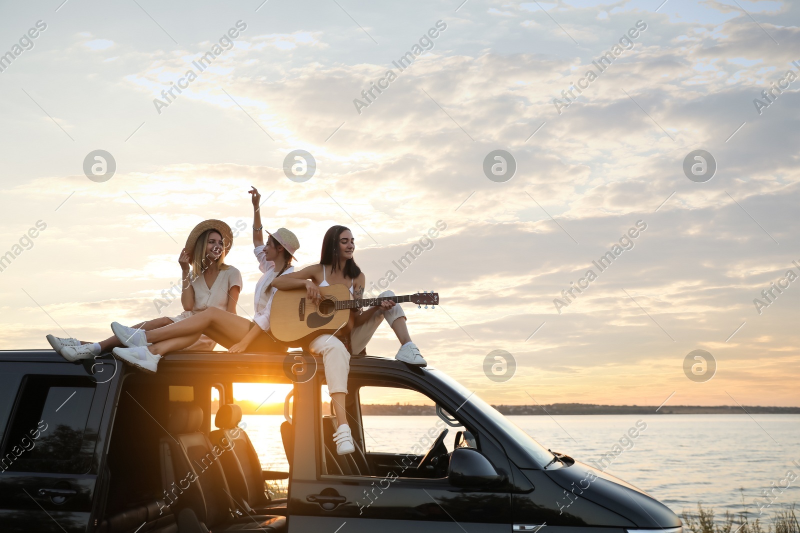 Happy friends with guitar sitting on car roof outdoors at sunset. Summer trip Photo of Happy friends with guitar sitting on car roof outdoors at sunset. Summer trip