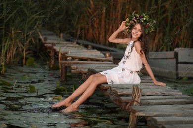 Cute little girl wearing wreath made of beautiful flowers on pier near pond at sunset Photo of Cute little girl wearing wreath made of beautiful flowers on pier near pond at sunset