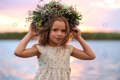 Photo of Cute little girl wearing wreath made of beautiful flowers near river at sunset