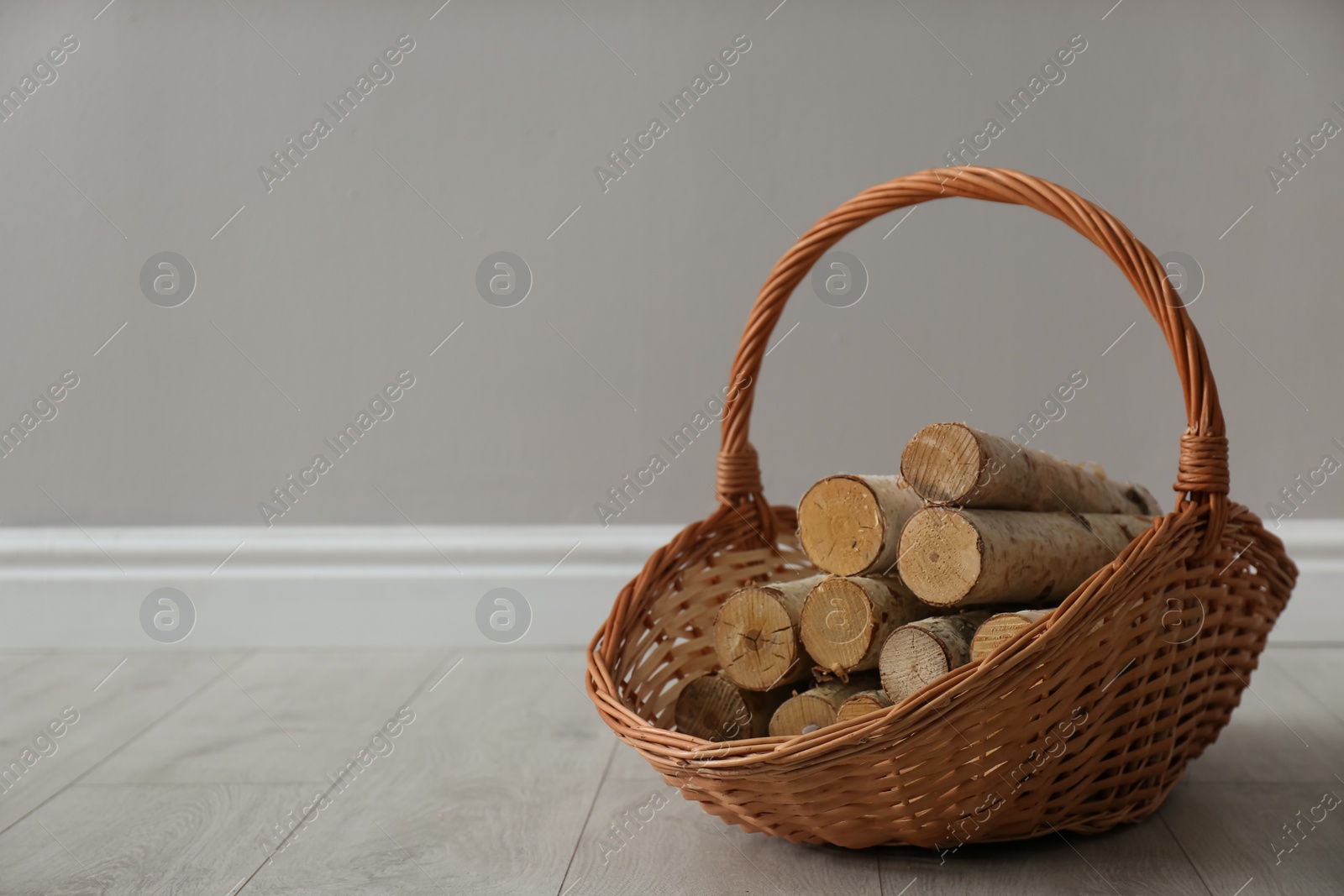 Wicker basket with firewood near grey wall indoors, space for text Photo of Wicker basket with firewood near grey wall indoors, space for text