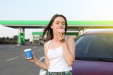 Beautiful young woman with coffee eating doughnut near car at gas station Photo of Beautiful young woman with coffee eating doughnut near car at gas station