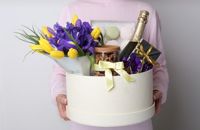 Woman holding basket with gifts, bouquet and champagne on light grey background, closeup Photo of Woman holding basket with gifts, bouquet and champagne on light grey background, closeup