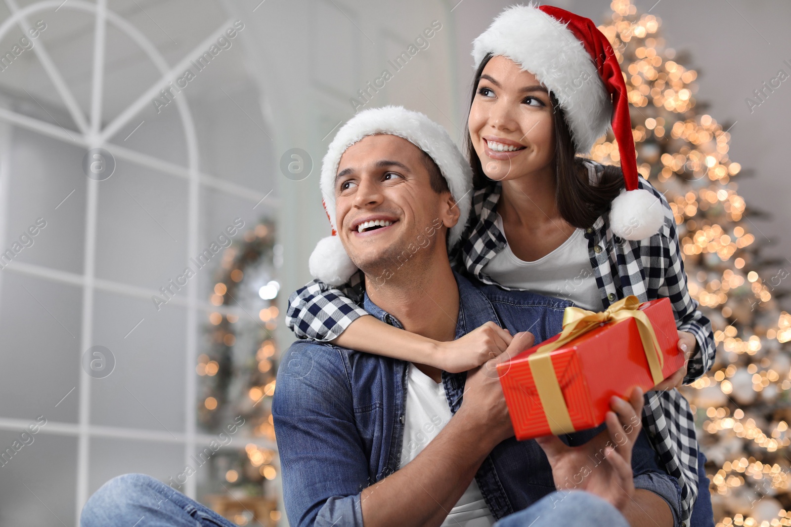 Happy young couple in Santa hats with Christmas gift at home Photo of Happy young couple in Santa hats with Christmas gift at home
