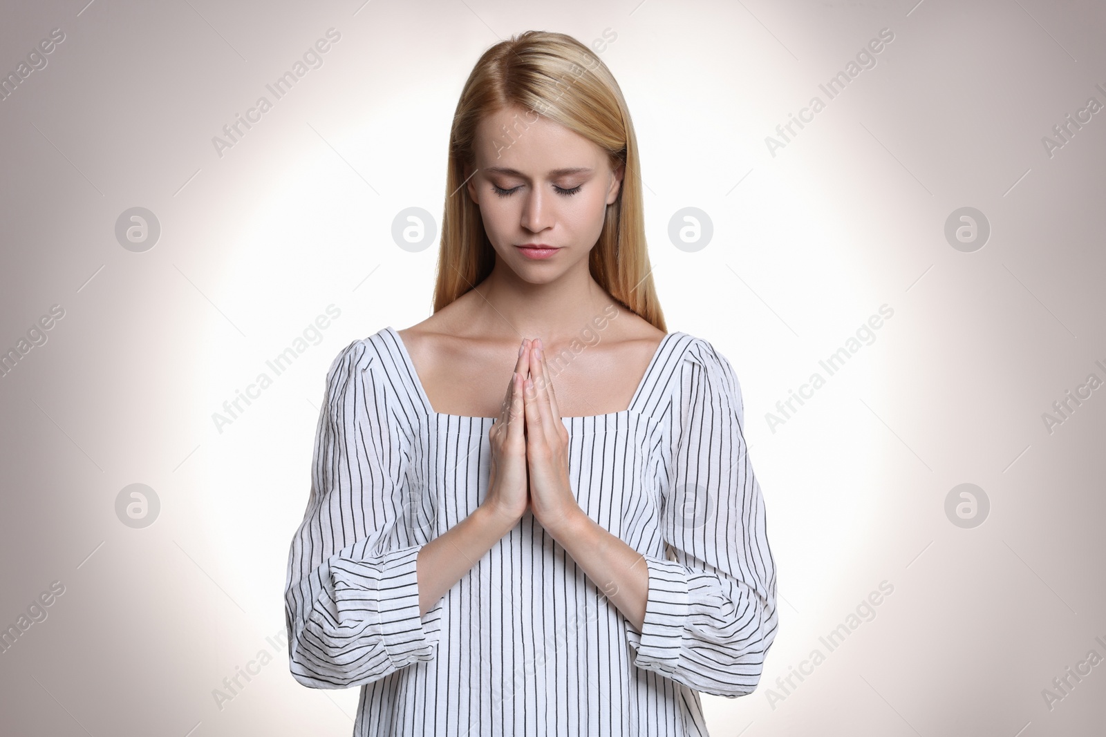 Religious young woman with clasped hands praying against light background Photo of Religious young woman with clasped hands praying against light background