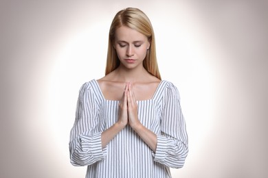 Religious young woman with clasped hands praying against light background Photo of Religious young woman with clasped hands praying against light background