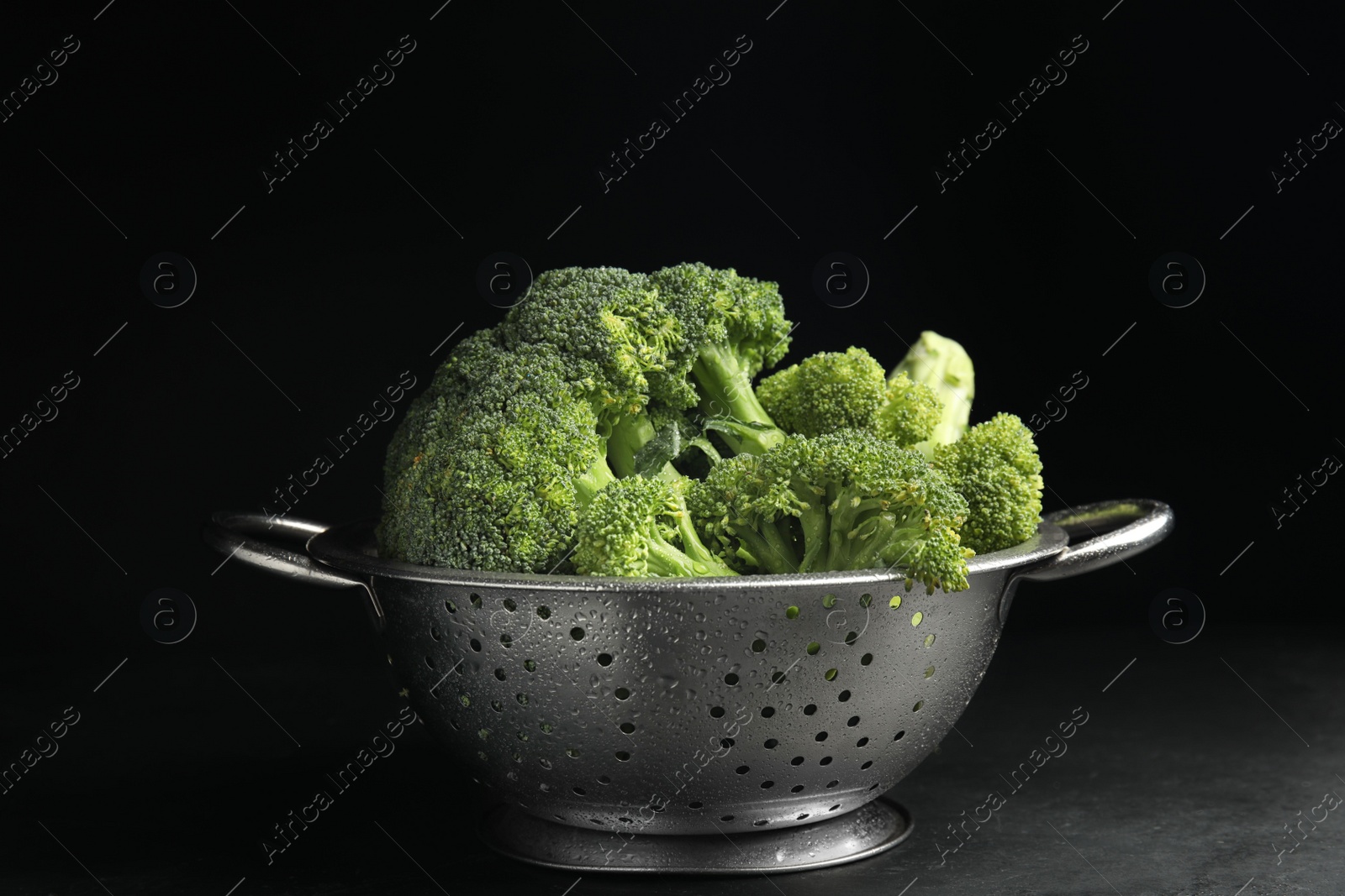 Tasty fresh broccoli in colander on table against black background Photo of Tasty fresh broccoli in colander on table against black background