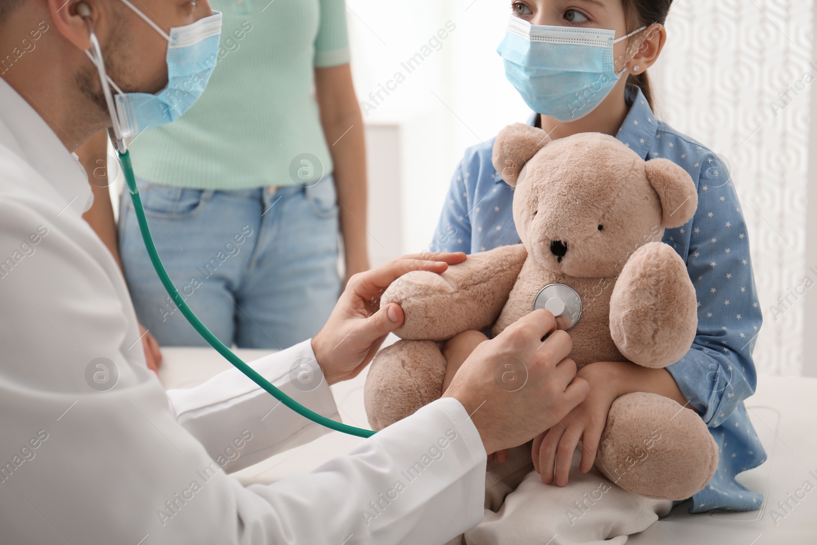 Pediatrician pretending to examine little girl's bear at hospital. Wearing protective masks Photo of Pediatrician pretending to examine little girl's bear at hospital. Wearing protective masks