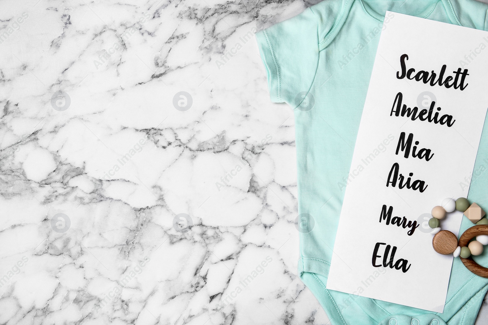 Bodysuit with list of baby names and toy on white marble background, top view. Space for text Photo of Bodysuit with list of baby names and toy on white marble background, top view. Space for text
