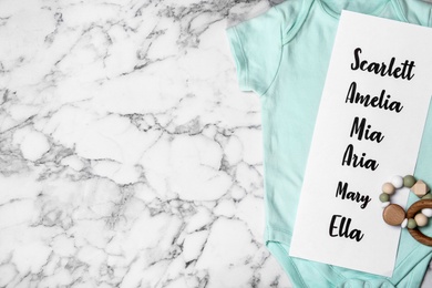 Bodysuit with list of baby names and toy on white marble background, top view. Space for text Photo of Bodysuit with list of baby names and toy on white marble background, top view. Space for text