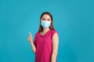 Vaccinated woman with protective mask and medical plaster on her arm showing thumb up against light blue background Photo of Vaccinated woman with protective mask and medical plaster on her arm showing thumb up against light blue background