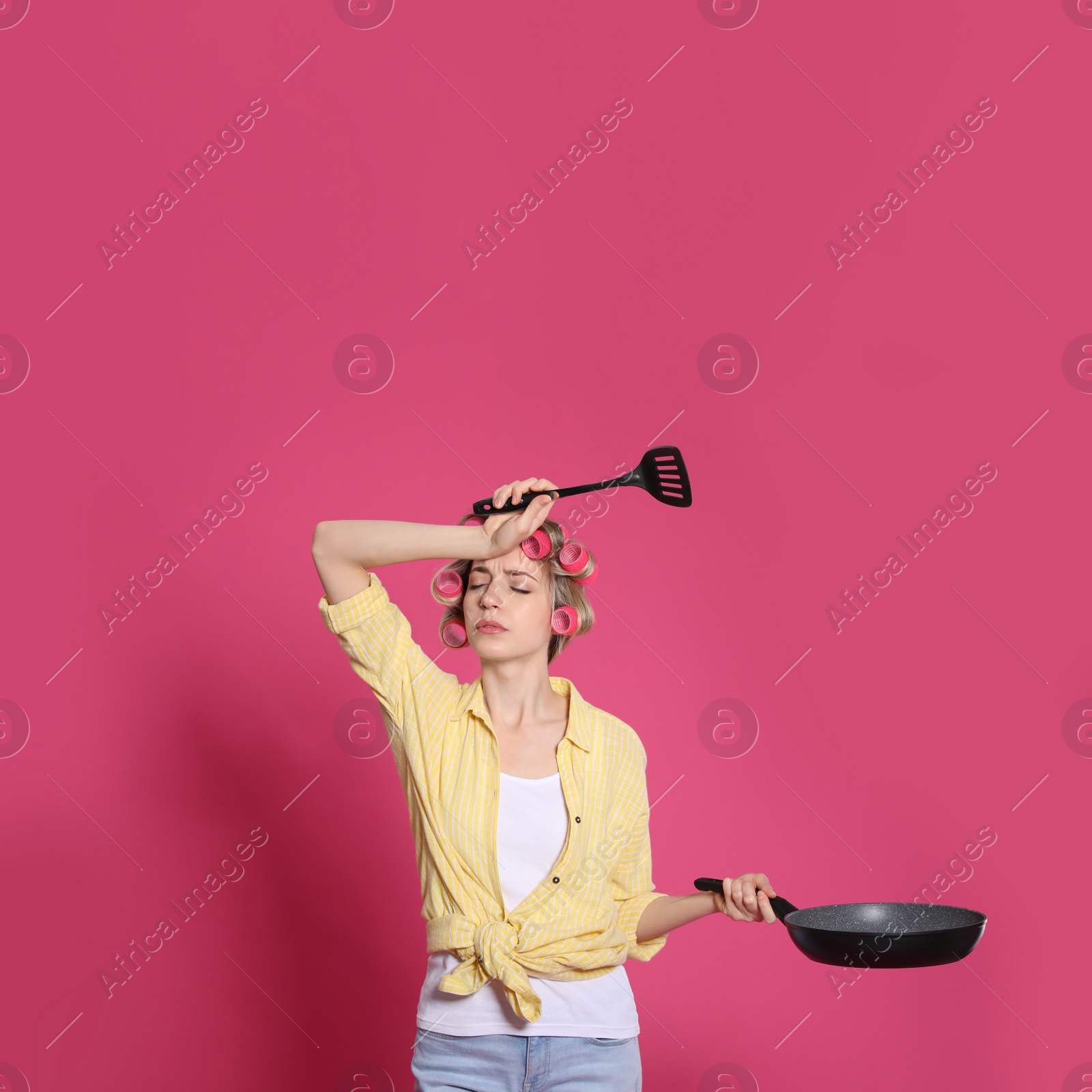 Young housewife with frying pan and spatula on pink background Photo of Young housewife with frying pan and spatula on pink background