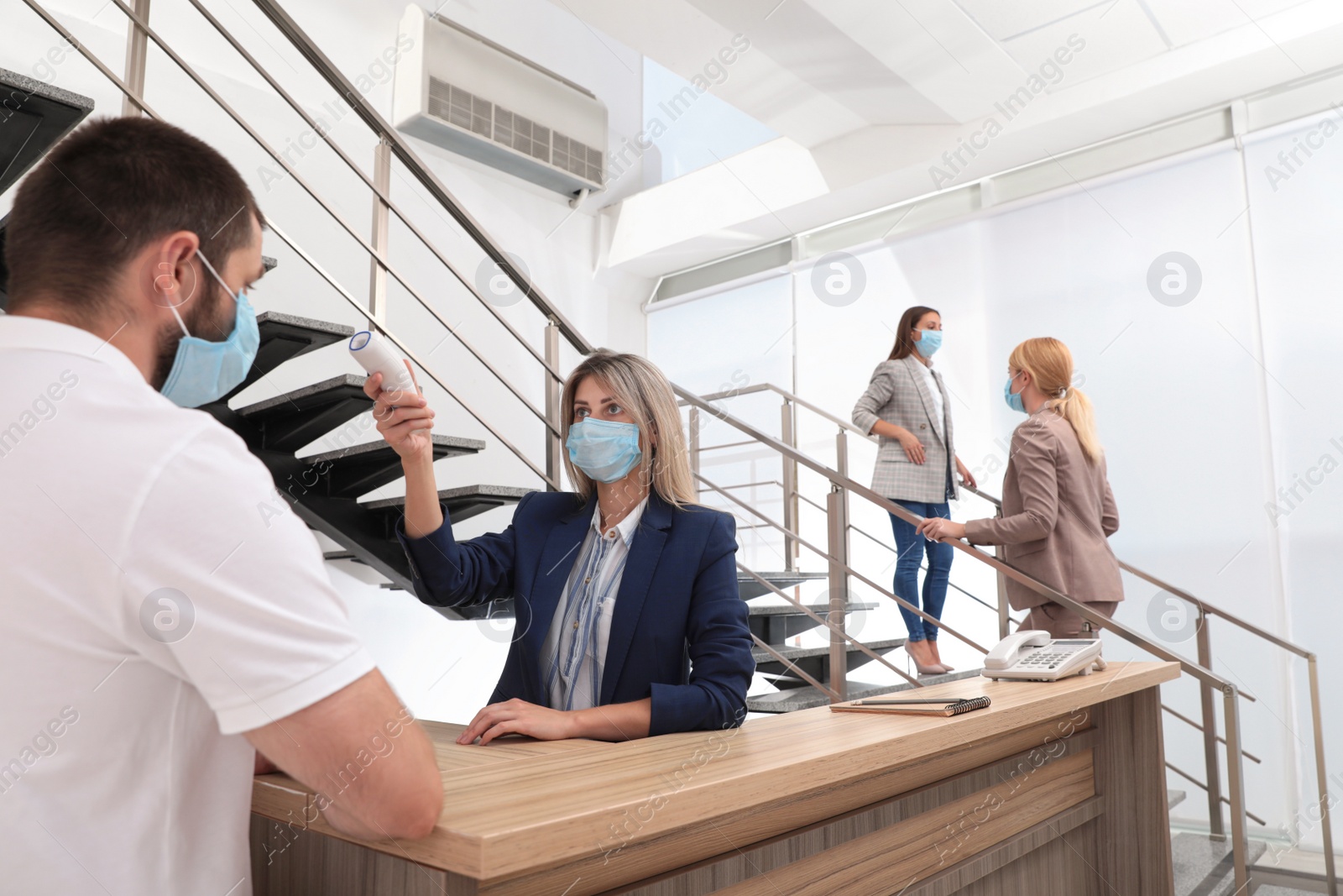 Woman in mask measuring temperature of employee with noncontact thermometer at office reception Photo of Woman in mask measuring temperature of employee with noncontact thermometer at office reception