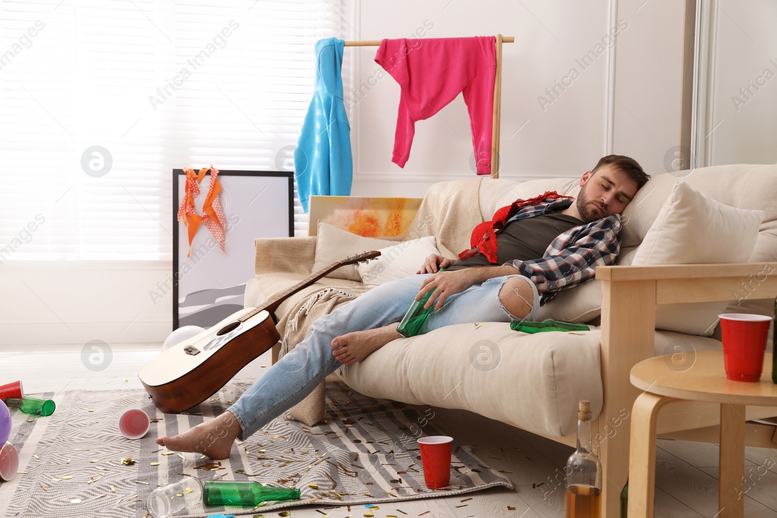 Young man with bottle of beer sleeping on sofa in messy room after party Photo of Young man with bottle of beer sleeping on sofa in messy room after party