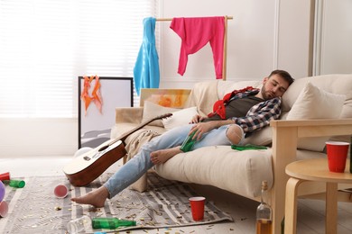 Young man with bottle of beer sleeping on sofa in messy room after party Photo of Young man with bottle of beer sleeping on sofa in messy room after party
