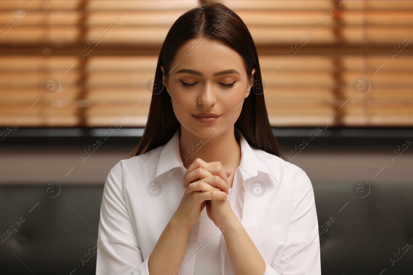 Religious young woman with clasped hands praying indoors Photo of Religious young woman with clasped hands praying indoors