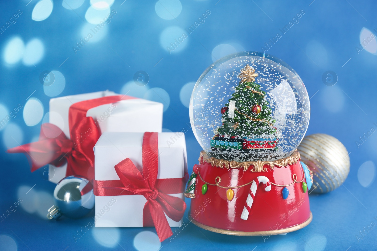 Beautiful Christmas snow globe, gift boxes and ornaments on blue background, bokeh effect Image of Beautiful Christmas snow globe, gift boxes and ornaments on blue background, bokeh effect