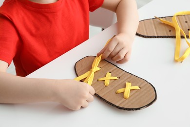 Photo of Little boy tying shoe lace using training cardboard template at white table, closeup