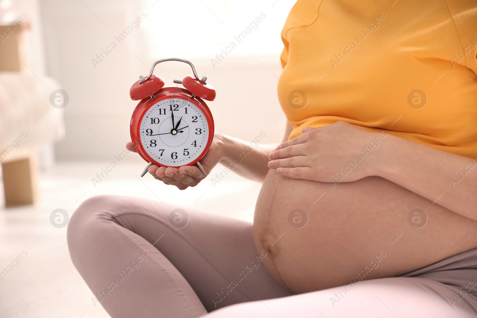 Young pregnant woman holding alarm clock near her belly at home, closeup. Time to give birth Photo of Young pregnant woman holding alarm clock near her belly at home, closeup. Time to give birth