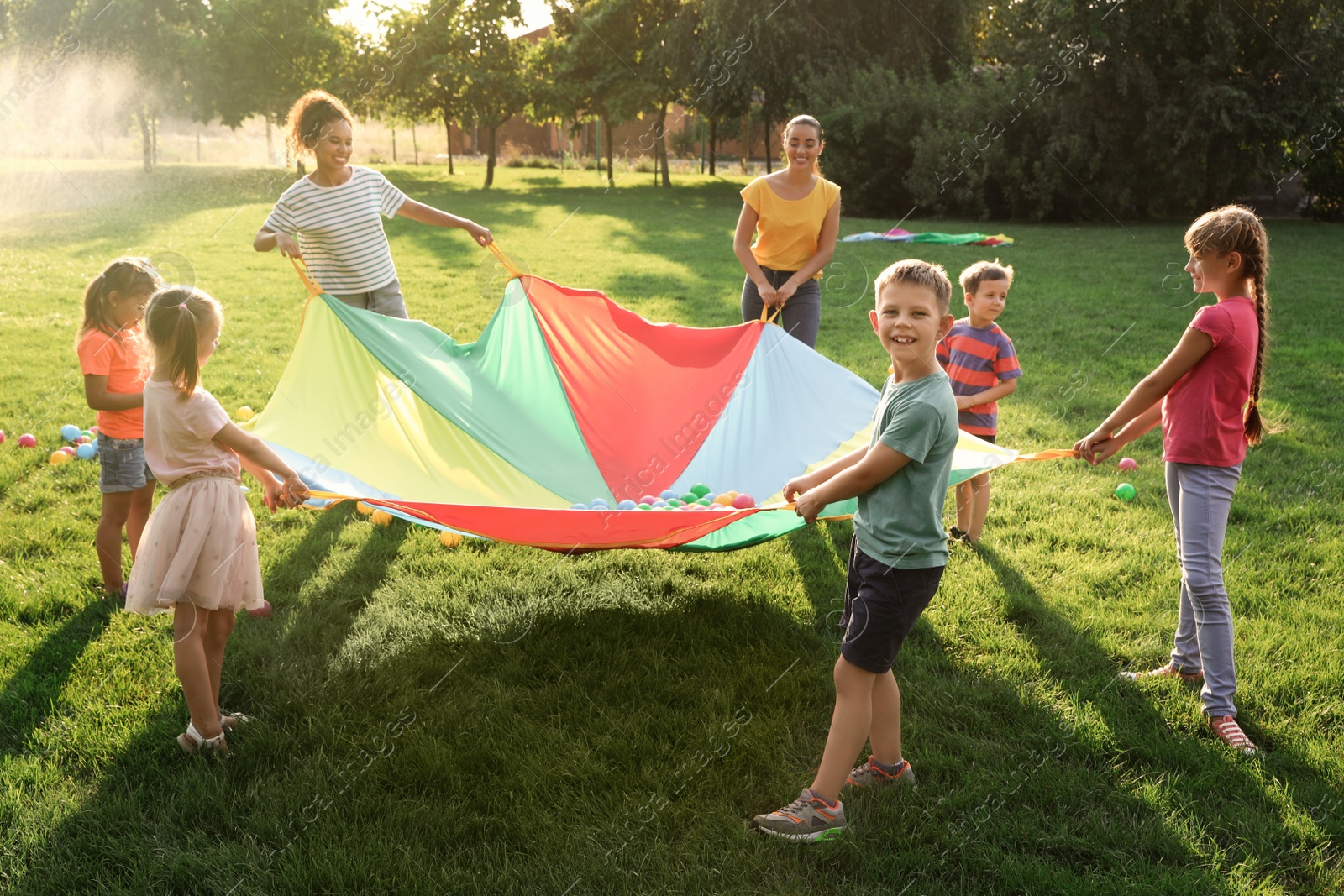 Group of children and teachers playing with rainbow playground parachute on green grass. Summer camp activity Photo of Group of children and teachers playing with rainbow playground parachute on green grass. Summer camp activity