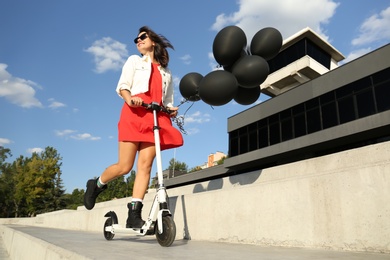 Young woman with black air balloons riding kick scooter along city street Photo of Young woman with black air balloons riding kick scooter along city street