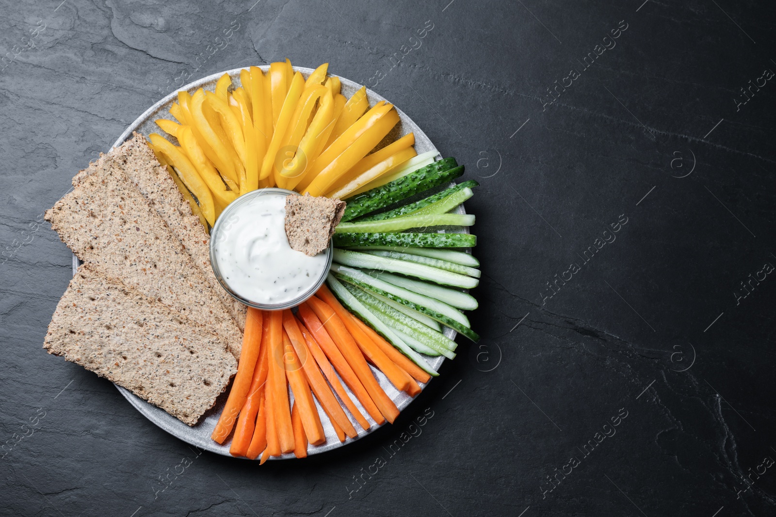 Plate with crispbreads, vegetable sticks and dip sauce on black table, top view. Space for text Photo of Plate with crispbreads, vegetable sticks and dip sauce on black table, top view. Space for text