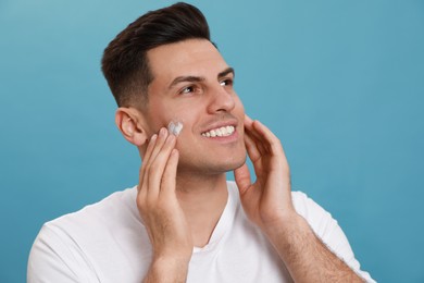 Happy handsome man applying face cream against turquoise background Photo of Happy handsome man applying face cream against turquoise background