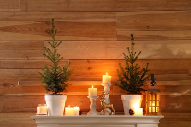 Small potted firs, candles and decor elements on white mantel near wooden wall Photo of Small potted firs, candles and decor elements on white mantel near wooden wall