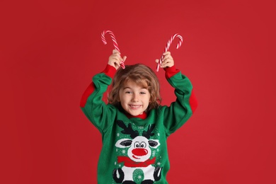 Cute little girl in Christmas sweater holding sweet candy canes near head against red background Photo of Cute little girl in Christmas sweater holding sweet candy canes near head against red background
