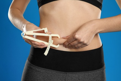 Young woman measuring body fat with caliper on blue background, closeup Photo of Young woman measuring body fat with caliper on blue background, closeup