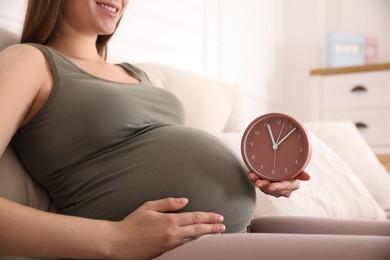 Young pregnant woman holding clock near her belly at home, closeup. Time to give birth Photo of Young pregnant woman holding clock near her belly at home, closeup. Time to give birth