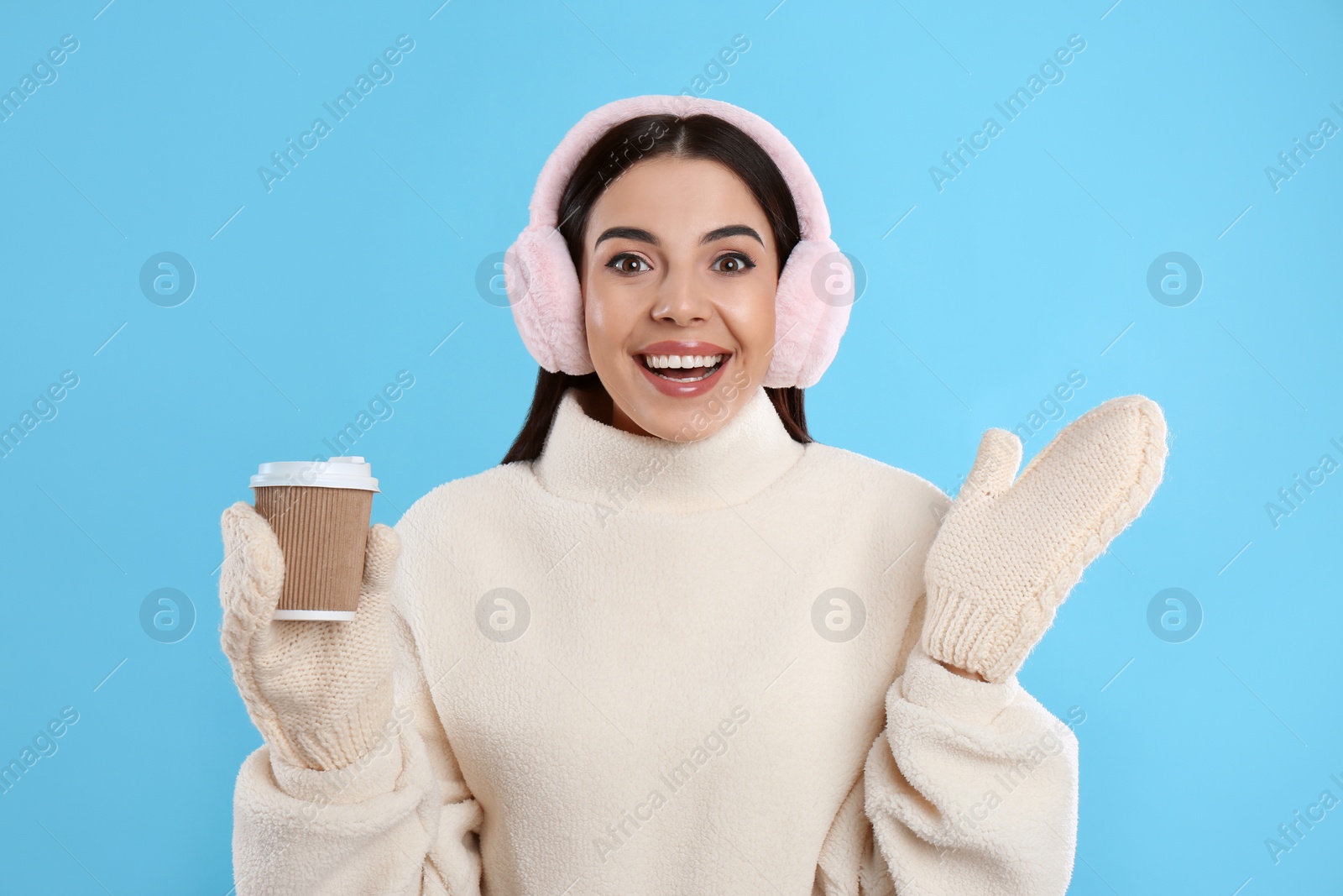 Beautiful young woman in earmuffs with cup of drink on light blue background Photo of Beautiful young woman in earmuffs with cup of drink on light blue background