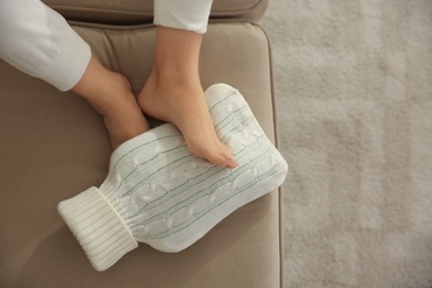 Woman warming feet with hot water bottle on sofa, closeup Photo of Woman warming feet with hot water bottle on sofa, closeup