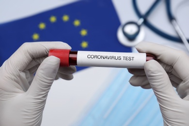 Doctor holding sample tube with label Coronavirus Test above medical items and European Union flag, closeup Photo of Doctor holding sample tube with label Coronavirus Test above medical items and European Union flag, closeup