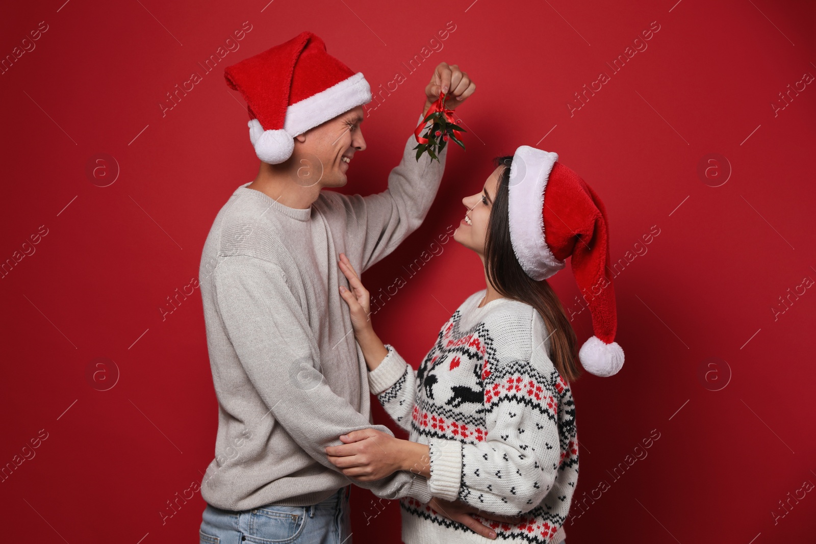 Happy couple in Santa hats standing under mistletoe bunch on red background Photo of Happy couple in Santa hats standing under mistletoe bunch on red background