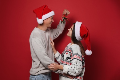 Happy couple in Santa hats standing under mistletoe bunch on red background Photo of Happy couple in Santa hats standing under mistletoe bunch on red background
